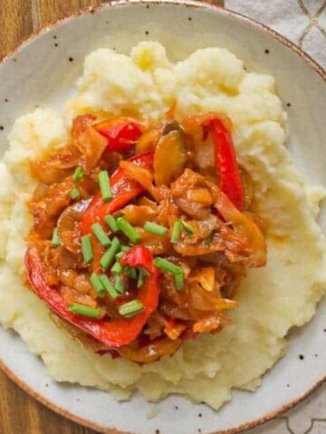 A plate of mashed potatoes topped with a sautéed vegetable mixture, including red bell peppers and onions, garnished with chopped chives.
