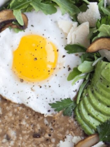 A bowl of oatmeal topped with a sunny-side-up egg, sliced avocado, arugula, mushrooms, parmesan shavings, and fresh herbs.