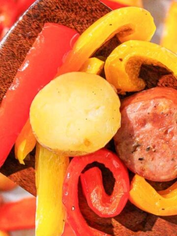 A wooden spatula holds sliced sausage, potatoes, and red and yellow bell peppers over a baking sheet with more of the same ingredients.