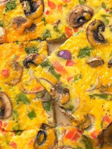 A baked egg casserole cut into squares, topped with sliced mushrooms, red and green peppers, red onions, and fresh herbs. A spatula lifts one piece from the tray.