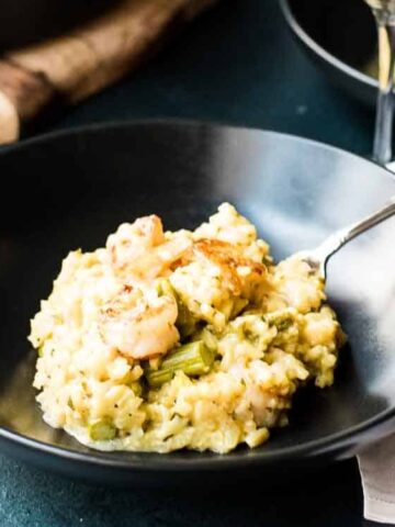 A black bowl contains creamy risotto with shrimp and vegetables, with a spoon resting inside the bowl on a dark surface.