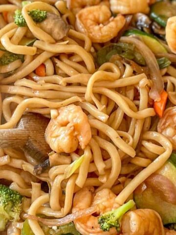 A wok filled with shrimp, udon noodles, and mixed vegetables being stirred with a wooden spoon.