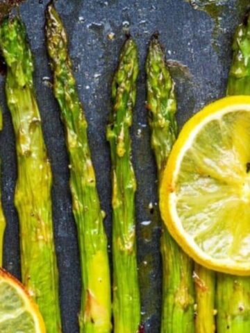 Roasted asparagus spears arranged in a row with two lemon slices on top, set on a dark, textured surface.