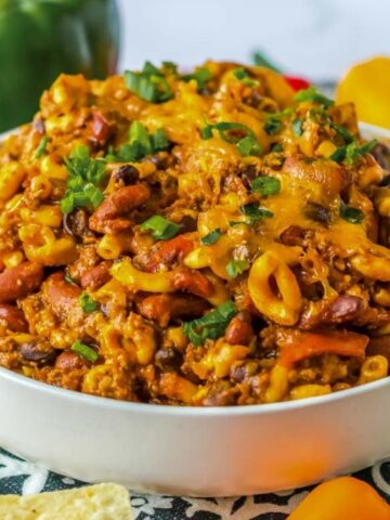 A bowl of chili mac topped with melted cheddar cheese and chopped green onions, surrounded by tortilla chips, bell peppers, and green onions.