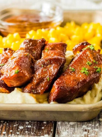 A tray with barbecued meat on top of mashed potatoes, served with a side of yellow corn and parsley garnish.