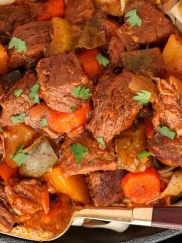 A close-up of a beef stew with carrots, potatoes, and bell peppers, served on top of wide noodles in a black dish, garnished with fresh herbs.
