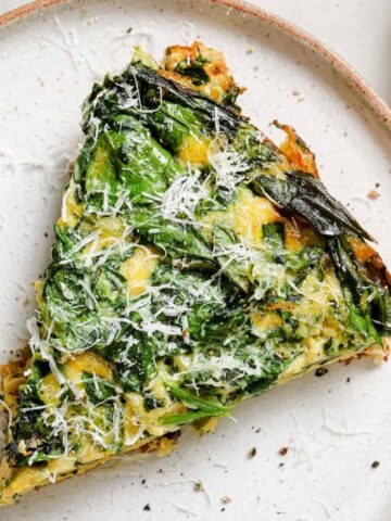 A slice of vegetable frittata topped with grated cheese is served on a white plate.