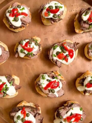 A round copper tray with 13 small crostini topped with sliced meat, cream, capers, and strips of red pepper, arranged in rows.