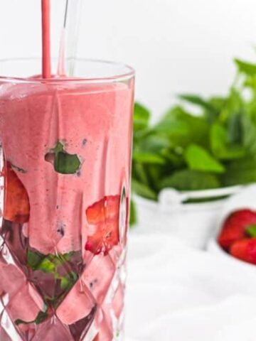 A thick pink smoothie is being poured into a glass with strawberries and a straw, with fresh strawberries and green leaves in the background.