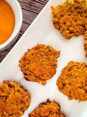 Six baked vegetable fritters are arranged on a white rectangular plate beside a bowl of orange dipping sauce.