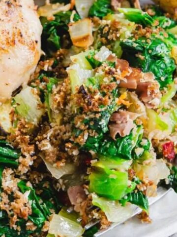 A plate with cooked chicken and a serving of sautéed leafy greens mixed with breadcrumbs and vegetables, with a fork on the side.