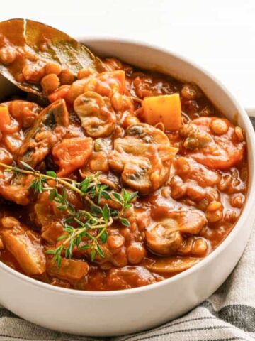 A bowl of vegetable stew with beans, carrots, mushrooms, and herbs, garnished with thyme and a bay leaf, on a striped cloth.