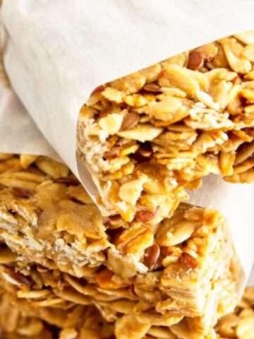 Two granola bars partially wrapped in parchment paper are stacked on a beige surface.