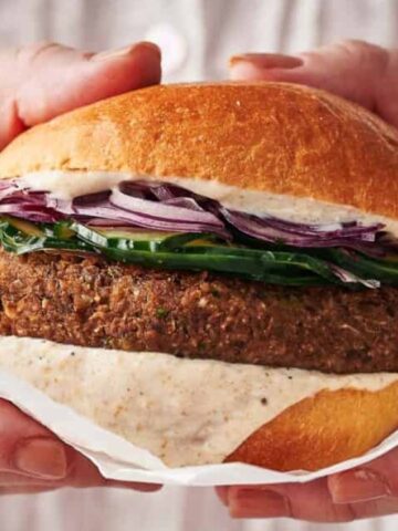 A person holding a plant-based burger with lettuce, onions, and sauce in a round bun wrapped in white paper.