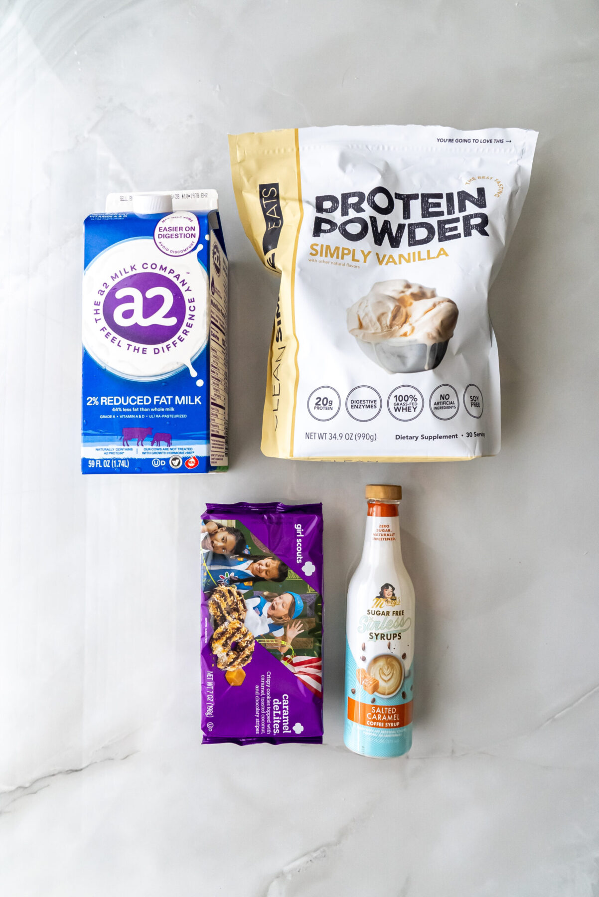 Flat lay of key ingredients: a2 Milk, Clean Simple Eats Vanilla Protein Powder, Girl Scouts Caramel Delites, and Mrs. Rogers Sugar-Free Salted Caramel Syrup.