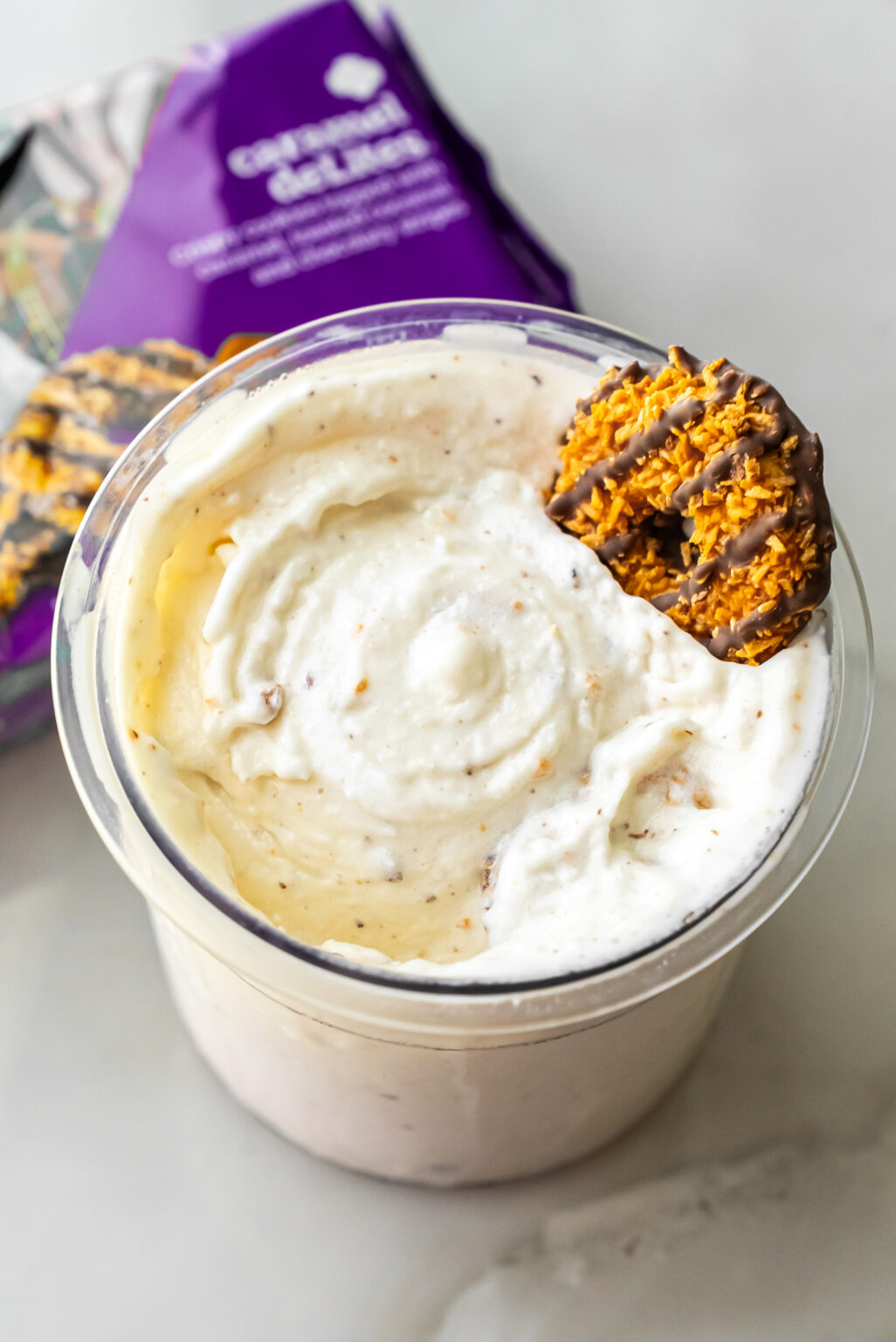 Close-up of Caramel DeLites Ninja Creami Ice Cream in a clear pint, topped with a Girl Scout cookie. A purple bag of Caramel deLites sits blurred in the background on a white surface.