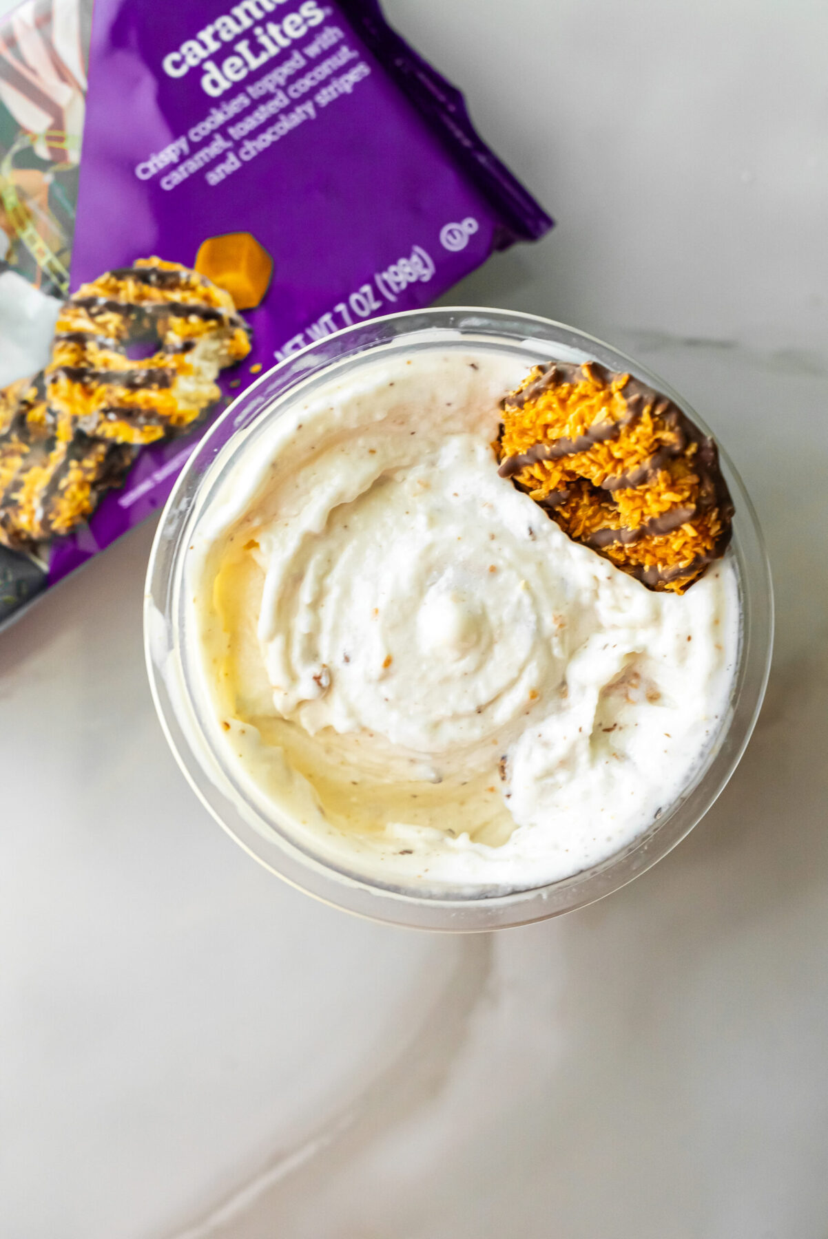 Overhead shot of creamy Caramel DeLites Ninja Creami Ice Cream in a pint, garnished with a coconut-caramel cookie. The purple packaging is visible in the upper left corner on a marble counter.