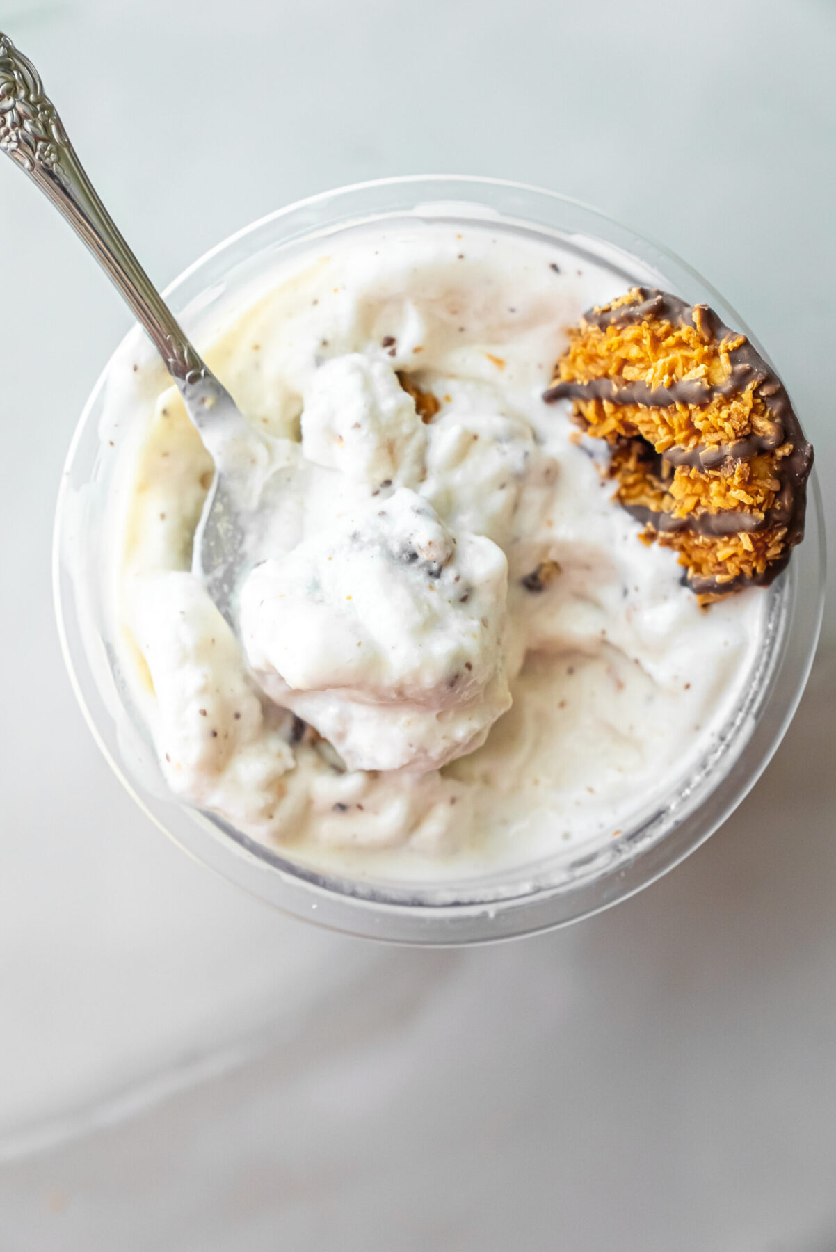 Top-down view of a silver spoon scooping Caramel DeLites Ninja Creami Ice Cream from a clear pint. A half-eaten cookie rests on the rim, showing the creamy texture mixed with cookie bits.