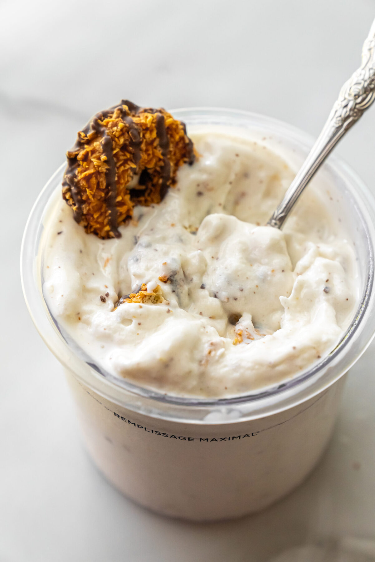Side view of Caramel DeLites Ninja Creami Ice Cream in a pint. A decorative spoon is inserted into the swirls, next to a chocolate-drizzled cookie garnish.