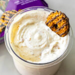 Close-up of Caramel DeLites Ninja Creami Ice Cream in a clear pint, topped with a Girl Scout cookie. A purple bag of Caramel deLites sits blurred in the background on a white surface.