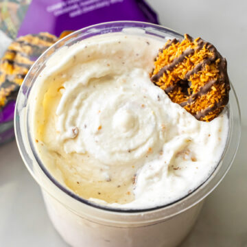 Close-up of Caramel DeLites Ninja Creami Ice Cream in a clear pint, topped with a Girl Scout cookie. A purple bag of Caramel deLites sits blurred in the background on a white surface.