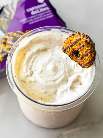 Close-up of Caramel DeLites Ninja Creami Ice Cream in a clear pint, topped with a Girl Scout cookie. A purple bag of Caramel deLites sits blurred in the background on a white surface.