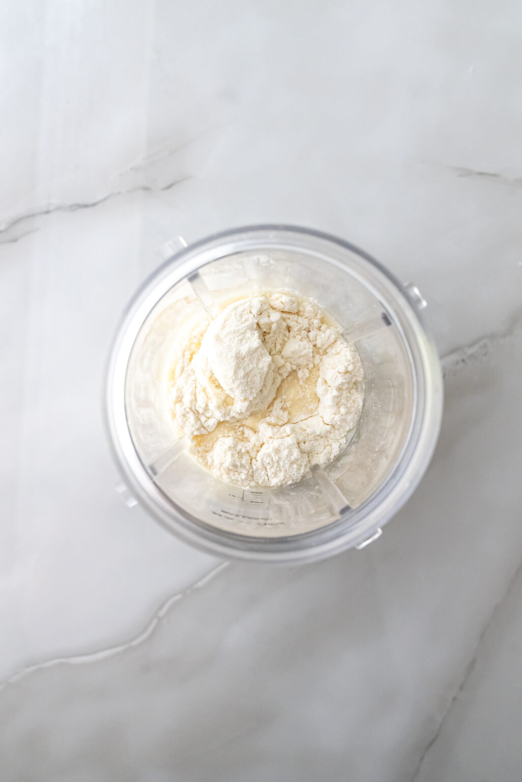 Top-down view of a clear blender cup on marble containing white protein powder added to a liquid base, preparing the mixture for Caramel DeLites Ninja Creami Ice Cream.