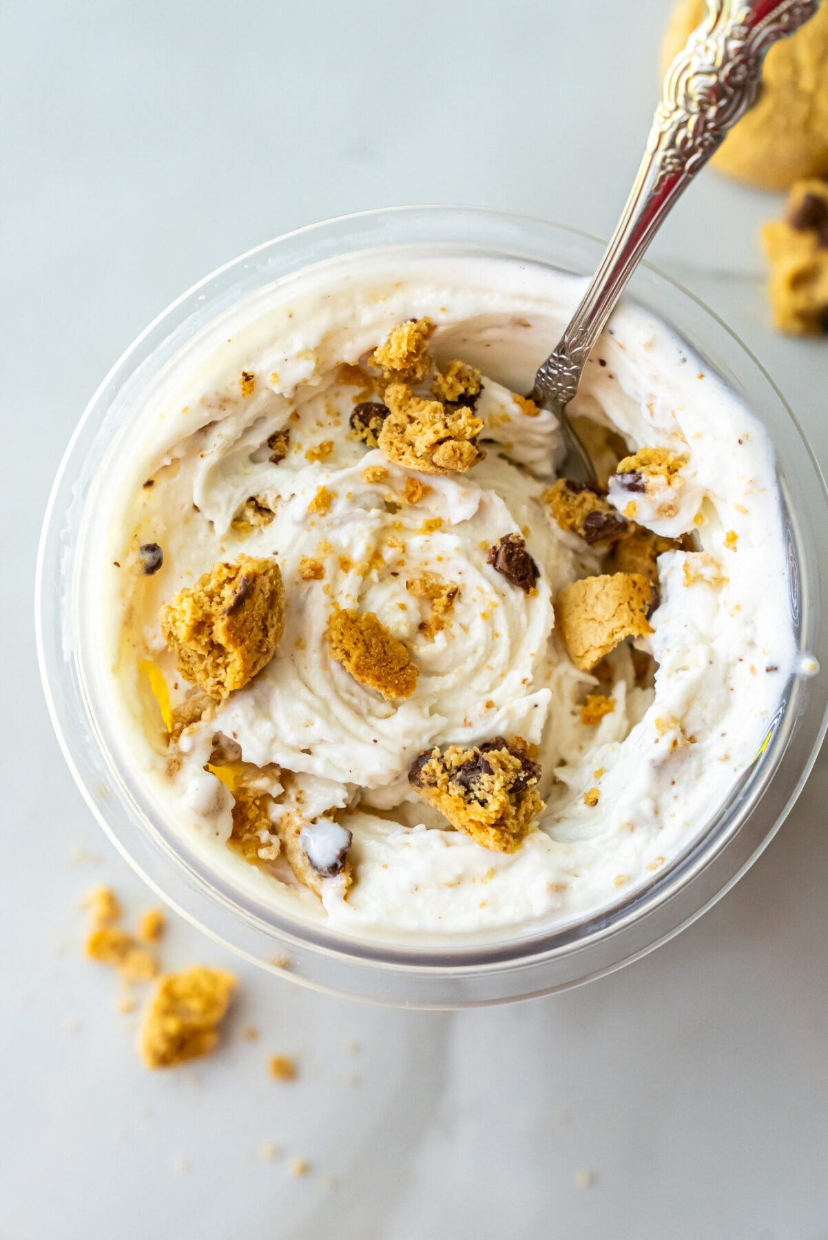 Overhead view of High Protein Caramel Chocolate Chip Ninja Creami Ice Cream in a clear pint. An ornate silver spoon is inserted into the white ice cream mixed with broken cookie chunks and chocolate chips.