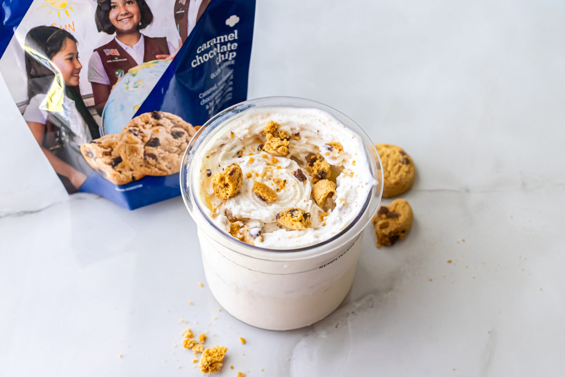 Close-up of High Protein Caramel Chocolate Chip Ninja Creami Ice Cream in a pint. Broken cookies top the swirls. A blue bag of Girl Scout Caramel Chocolate Chip cookies sits behind it.