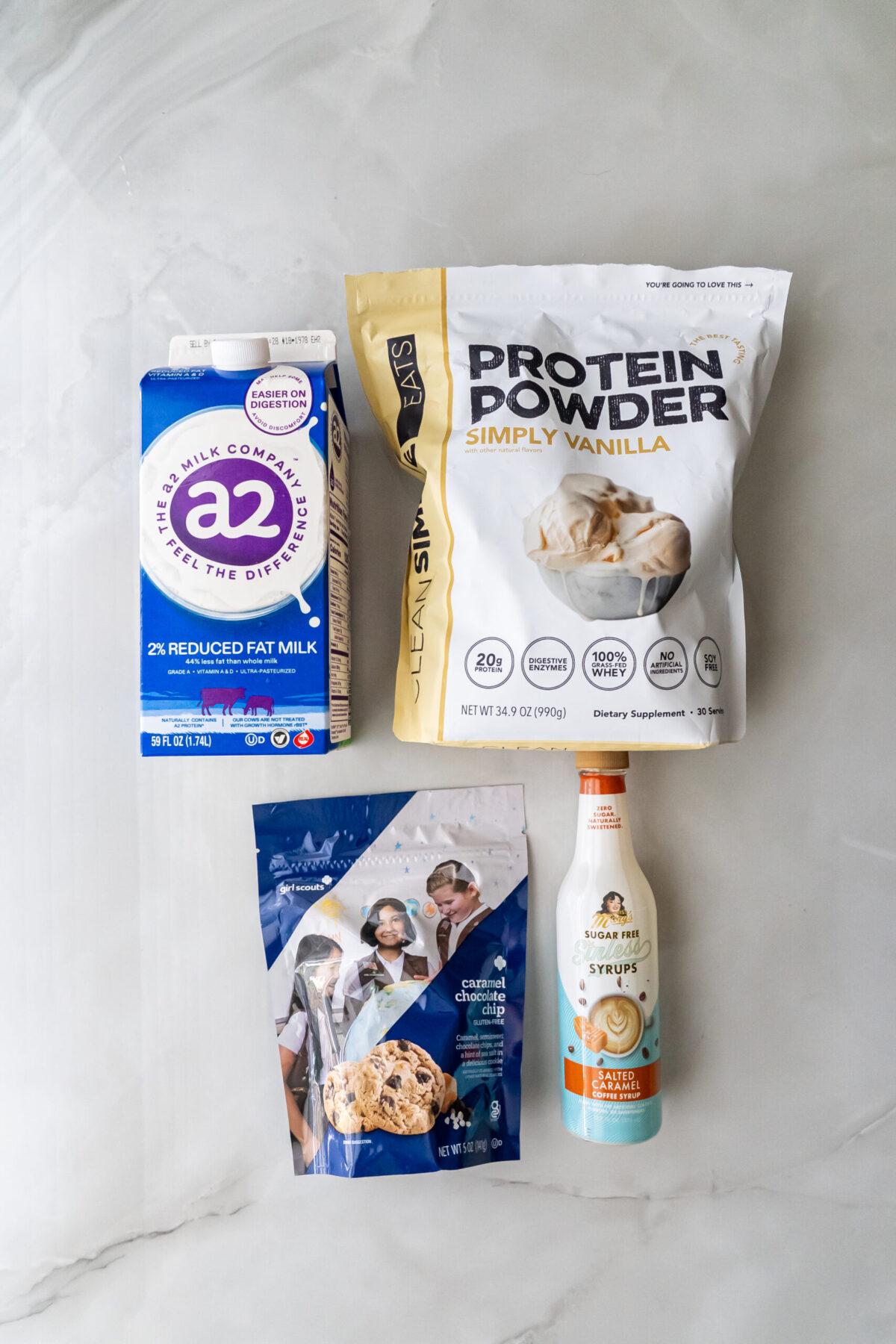 Flat lay of ingredients: a2 milk carton, Simply Vanilla protein powder bag, Girl Scout Caramel Chocolate Chip cookies package, and Mrs. Rogers Sugar Free Salted Caramel syrup on a white marble surface.
