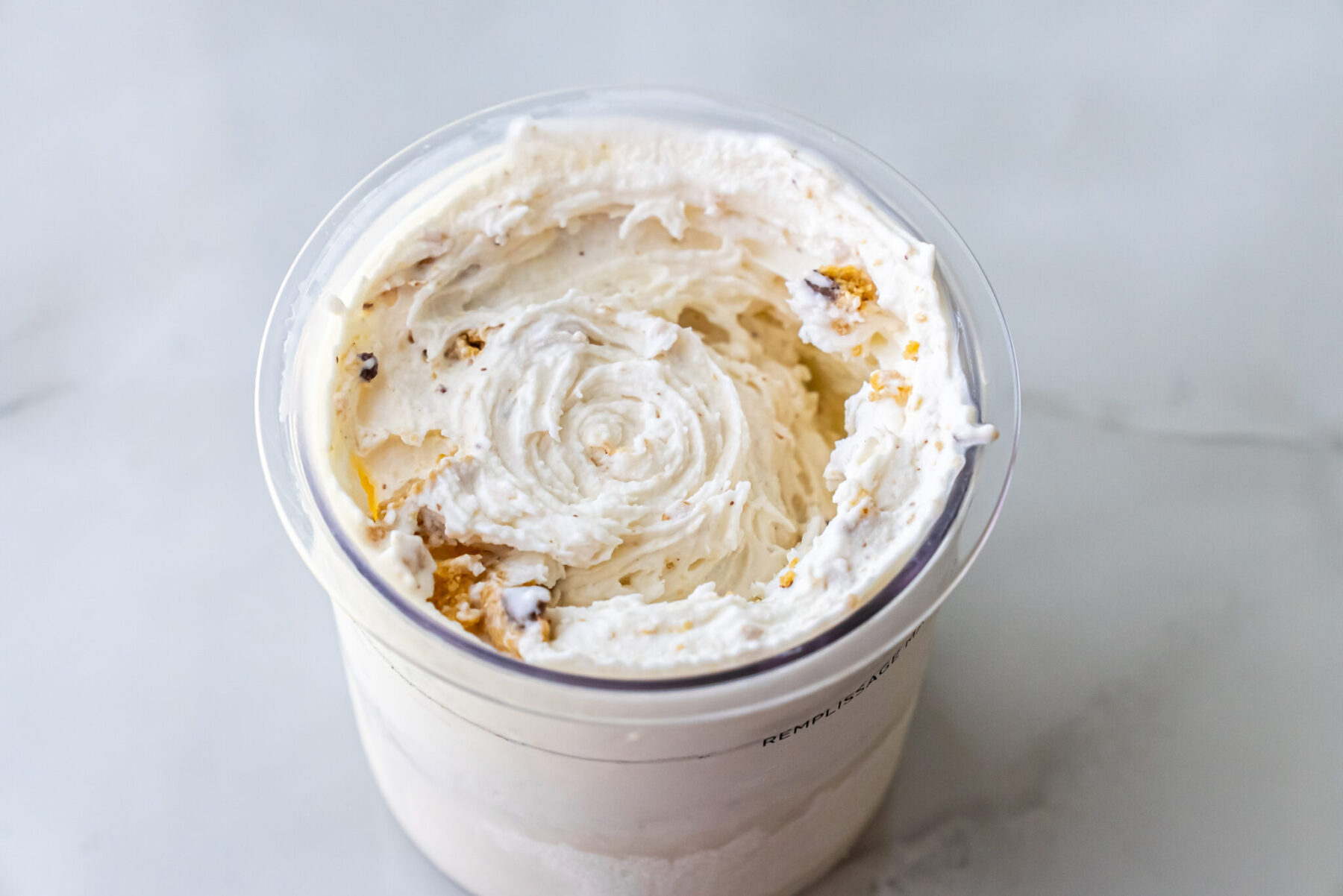 Top-down close-up of swirled High Protein Caramel Chocolate Chip Ninja Creami Ice Cream in a pint. Small pieces of caramel cookie and chocolate chips are visible throughout the creamy white base.