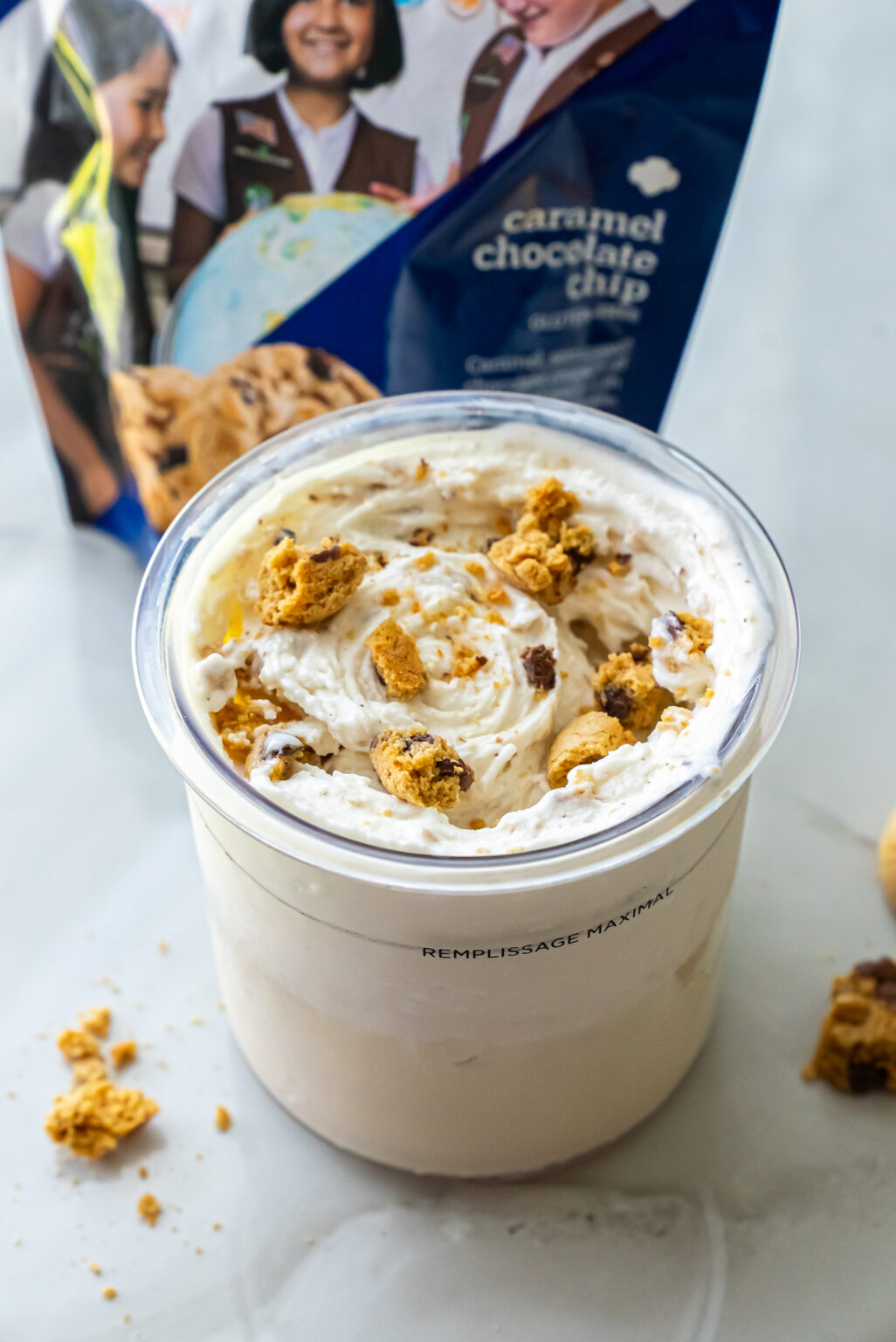Angled shot of High Protein Caramel Chocolate Chip Ninja Creami Ice Cream in a pint topped with broken cookie chunks. The blue Girl Scout Caramel Chocolate Chip cookie bag is blurred in the background.