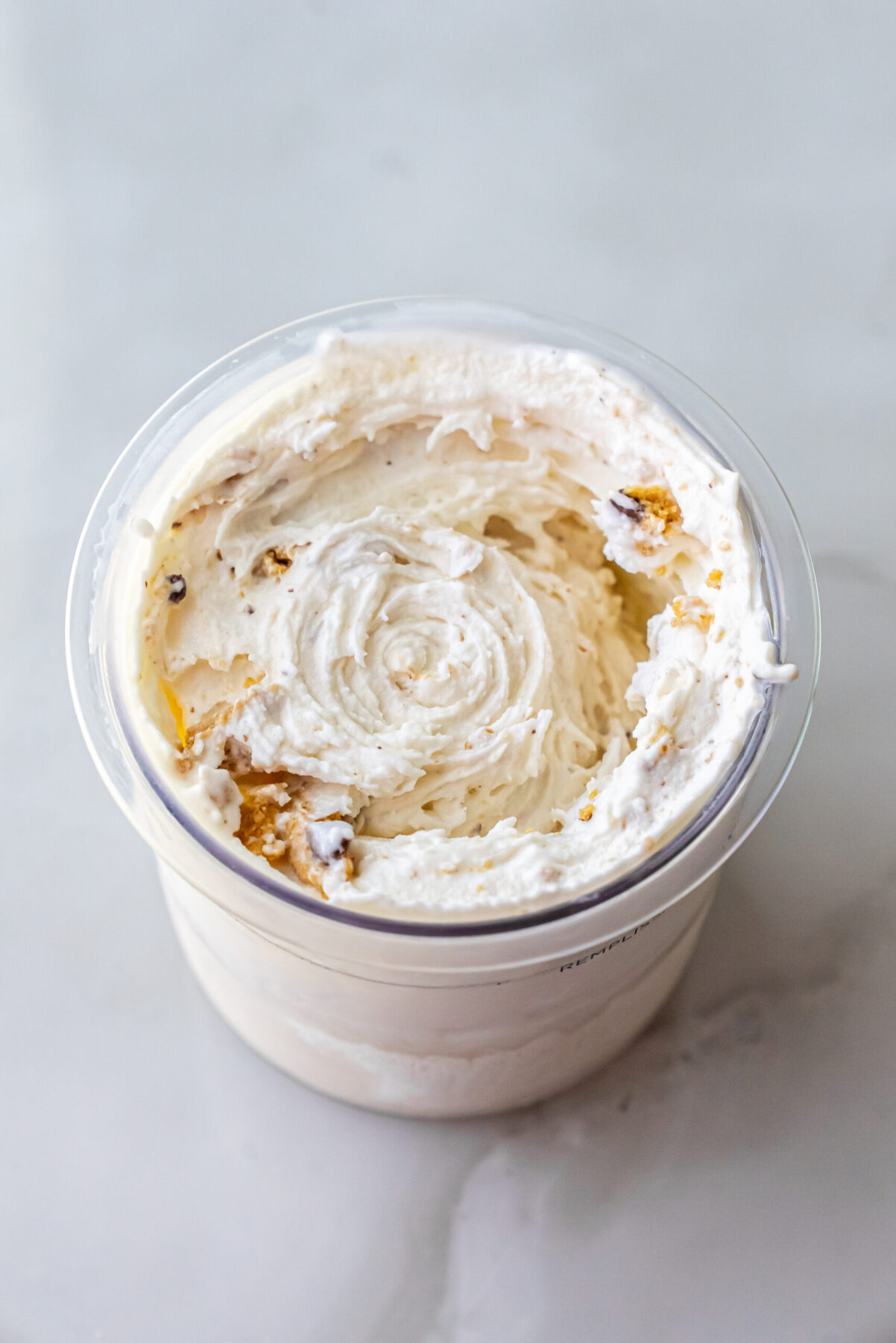 Direct top-down shot of swirled High Protein Caramel Chocolate Chip Ninja Creami Ice Cream in a clear pint. Small pieces of caramel cookie and chocolate chips are distributed throughout the creamy white base.