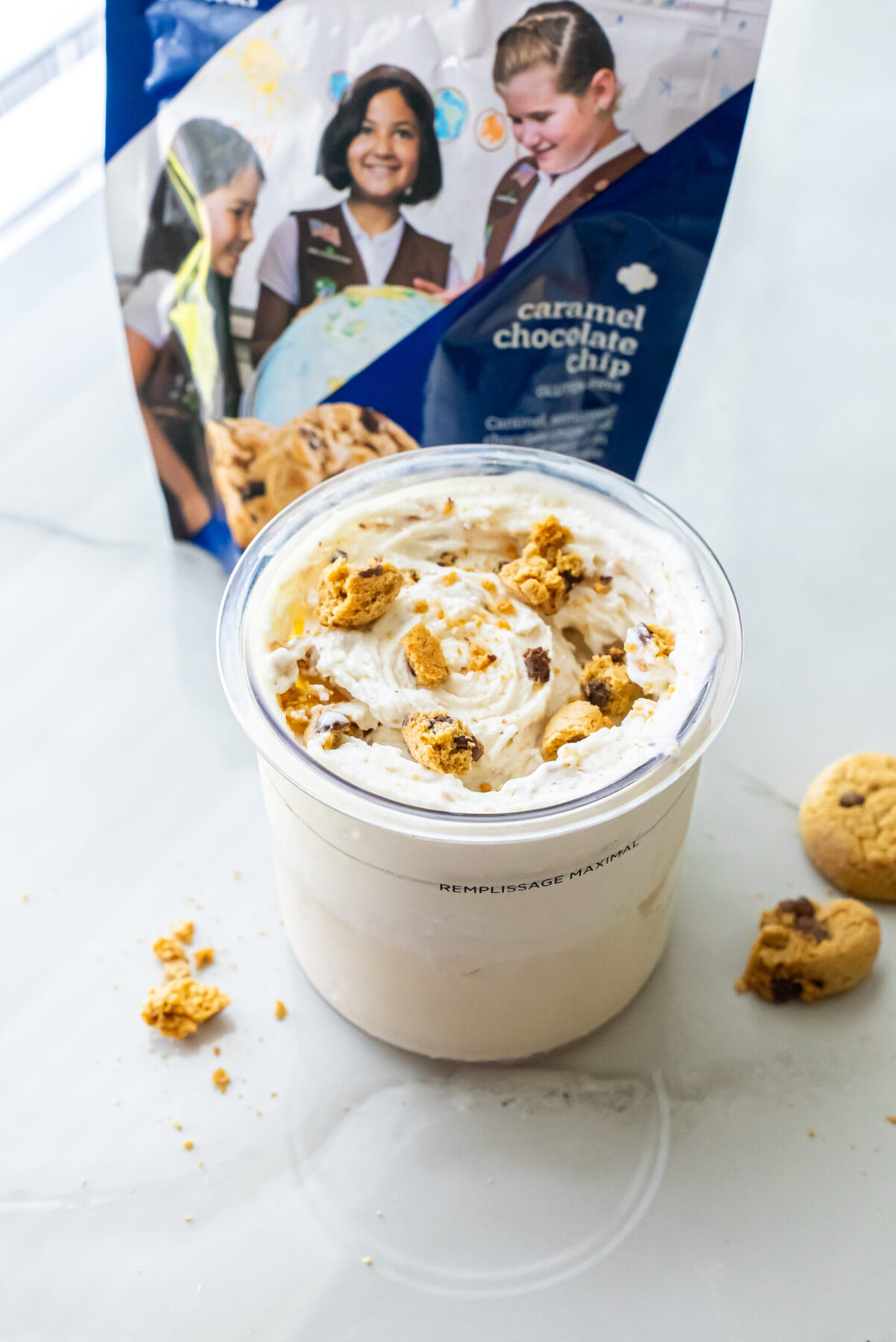Angled view of High Protein Caramel Chocolate Chip Ninja Creami Ice Cream in a pint topped with broken cookie chunks. The blue Girl Scout Caramel Chocolate Chip cookie bag stands behind it on a white marble counter.