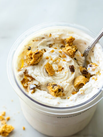 Close-up overhead view of High Protein Caramel Chocolate Chip Ninja Creami Ice Cream in a pint. A silver spoon rests in the white ice cream mixed with broken cookie chunks and chocolate chips. Crumbs scatter on the surface.