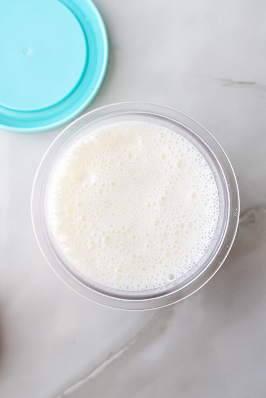 Top-down view of a clear Ninja Creami pint filled with frothy, bubbly white liquid base containing yellow syrup swirls, ready to be frozen for High Protein Caramel Chocolate Chip Ninja Creami Ice Cream. A teal lid sits nearby.