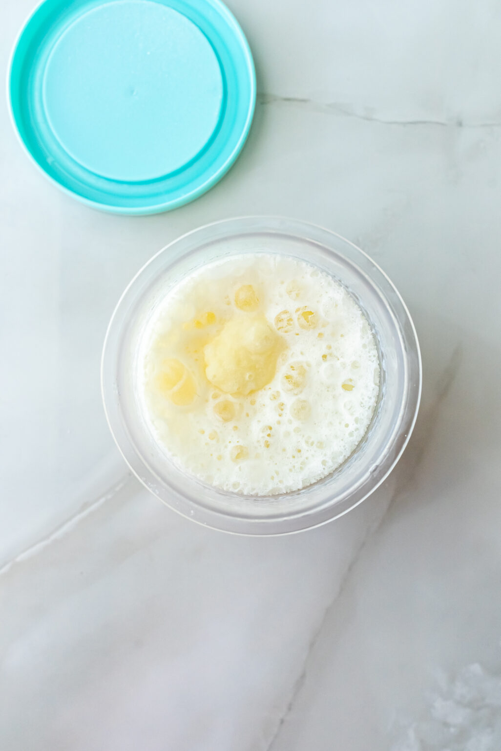 Overhead shot of smooth, uniformly frothed white liquid base in a clear Ninja Creami pint, prepared for freezing before processing into High Protein Caramel Chocolate Chip Ninja Creami Ice Cream. Teal lid visible in corner.