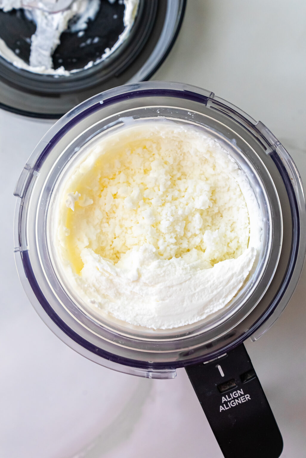 Top-down view of churned white ice cream in a Ninja Creami pint showing coarse, icy texture after initial spin cycle for High Protein Caramel Chocolate Chip Ninja Creami Ice Cream. Black machine lid rests beside it.