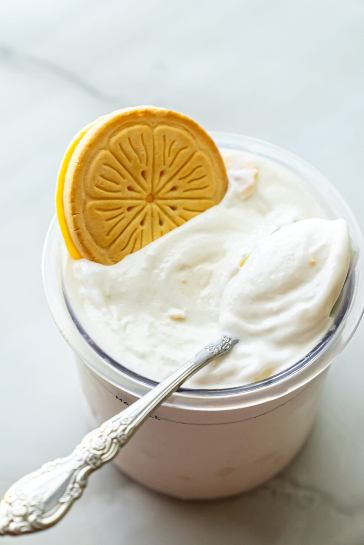 Close-up of Lemonades Ninja Creami Ice Cream in a clear pint. A whole Girl Scout Lemonades cookie stands upright in the white ice cream next to an ornate silver spoon resting on the rim.