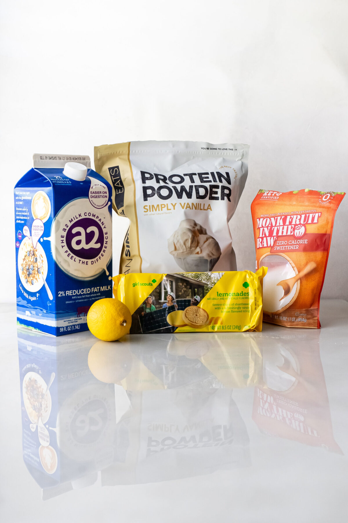 Flat lay of ingredients for Lemonades Ninja Creami Ice Cream: a2 milk carton, Simply Vanilla protein powder bag, fresh lemon, Girl Scout Lemonades package, and Monk Fruit sweetener on a reflective white surface.