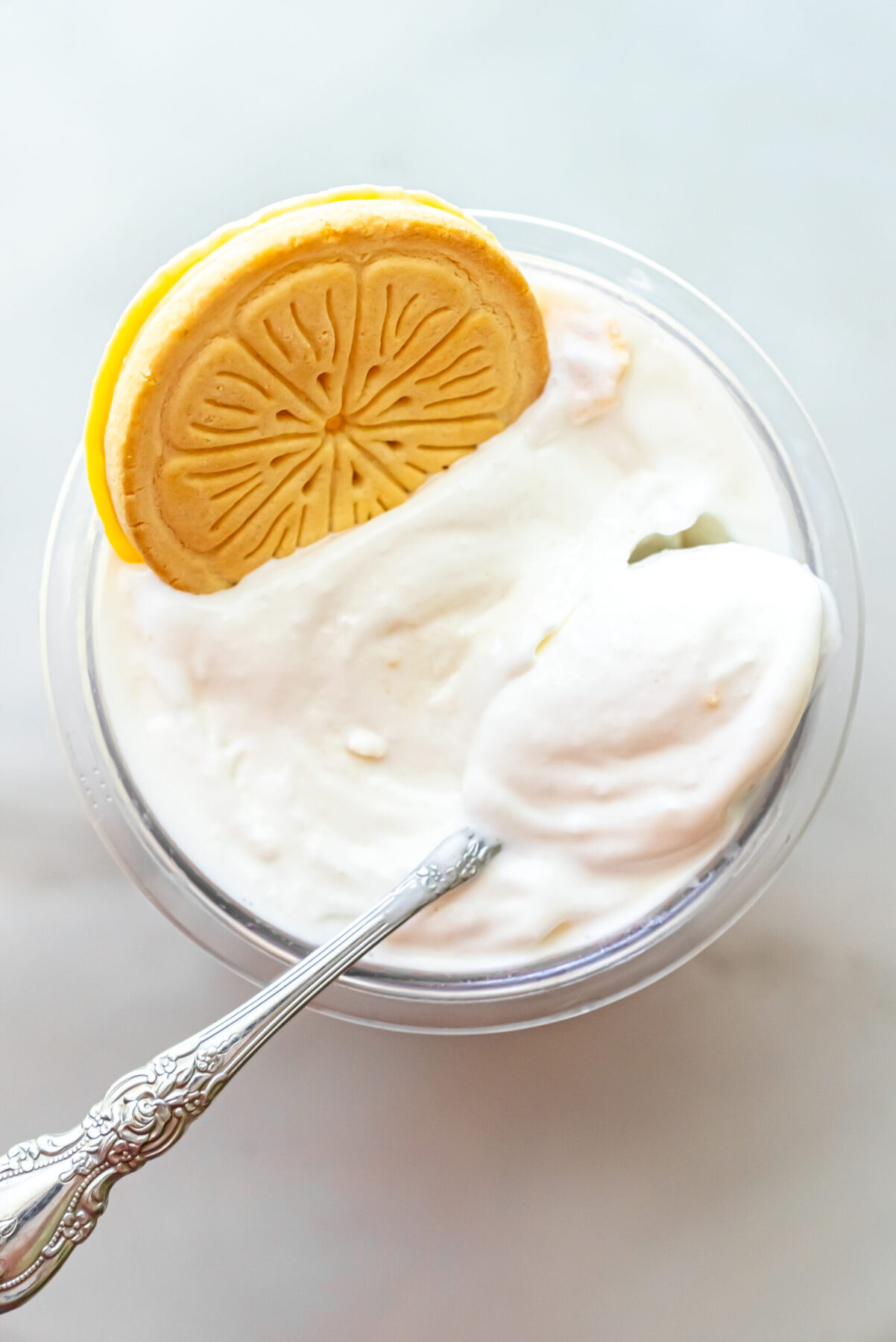 Overhead view of Lemonades Ninja Creami Ice Cream in a pint. An ornate silver spoon rests in the white ice cream, and a whole Lemonades cookie sits on the upper left edge of the container.