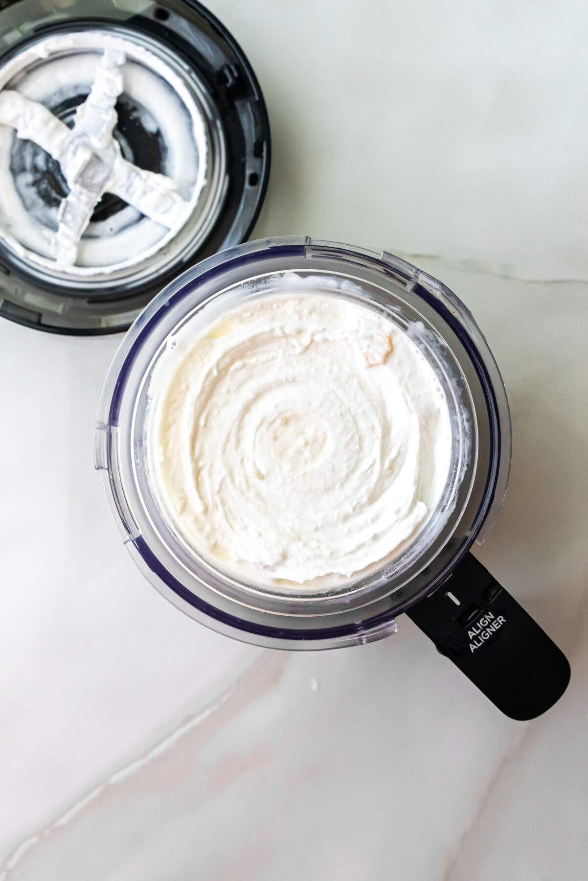 Top-down view of swirled white Lemonades Ninja Creami Ice Cream in a clear pint. The black machine lid with cream residue sits beside the container on a marble surface.