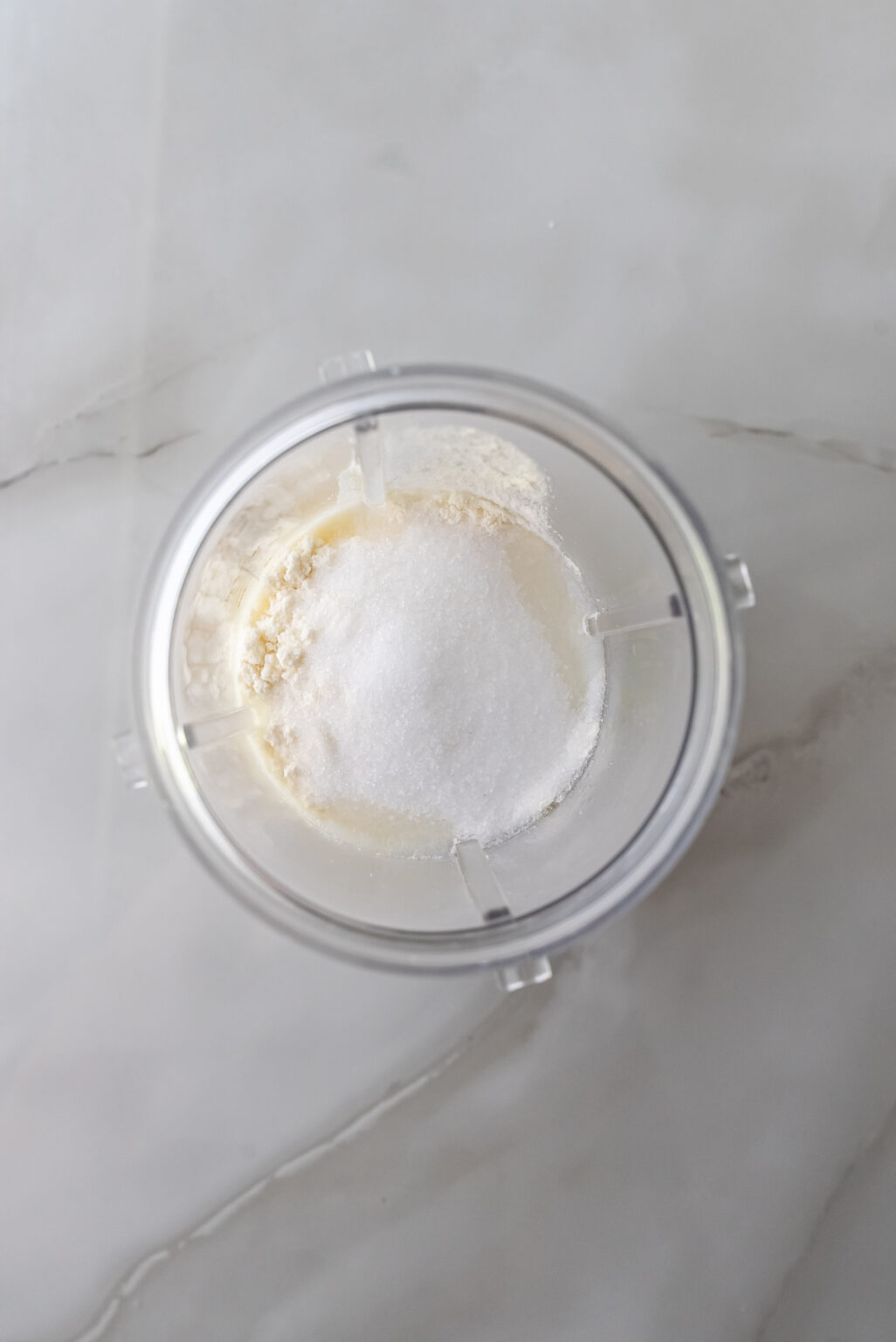 Top-down view of a clear Ninja Creami pint containing white protein powder and granulated sweetener over liquid base, preparing the mixture for Lemonades Ninja Creami Ice Cream.