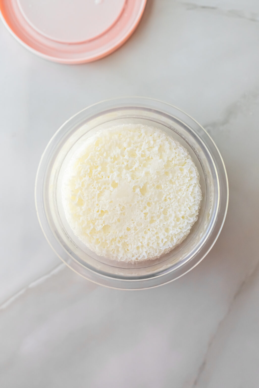 Overhead shot of the frozen, porous white ice cream base inside a clear Ninja Creami pint with a pink lid nearby, ready to be processed into Lemonades Ninja Creami Ice Cream.