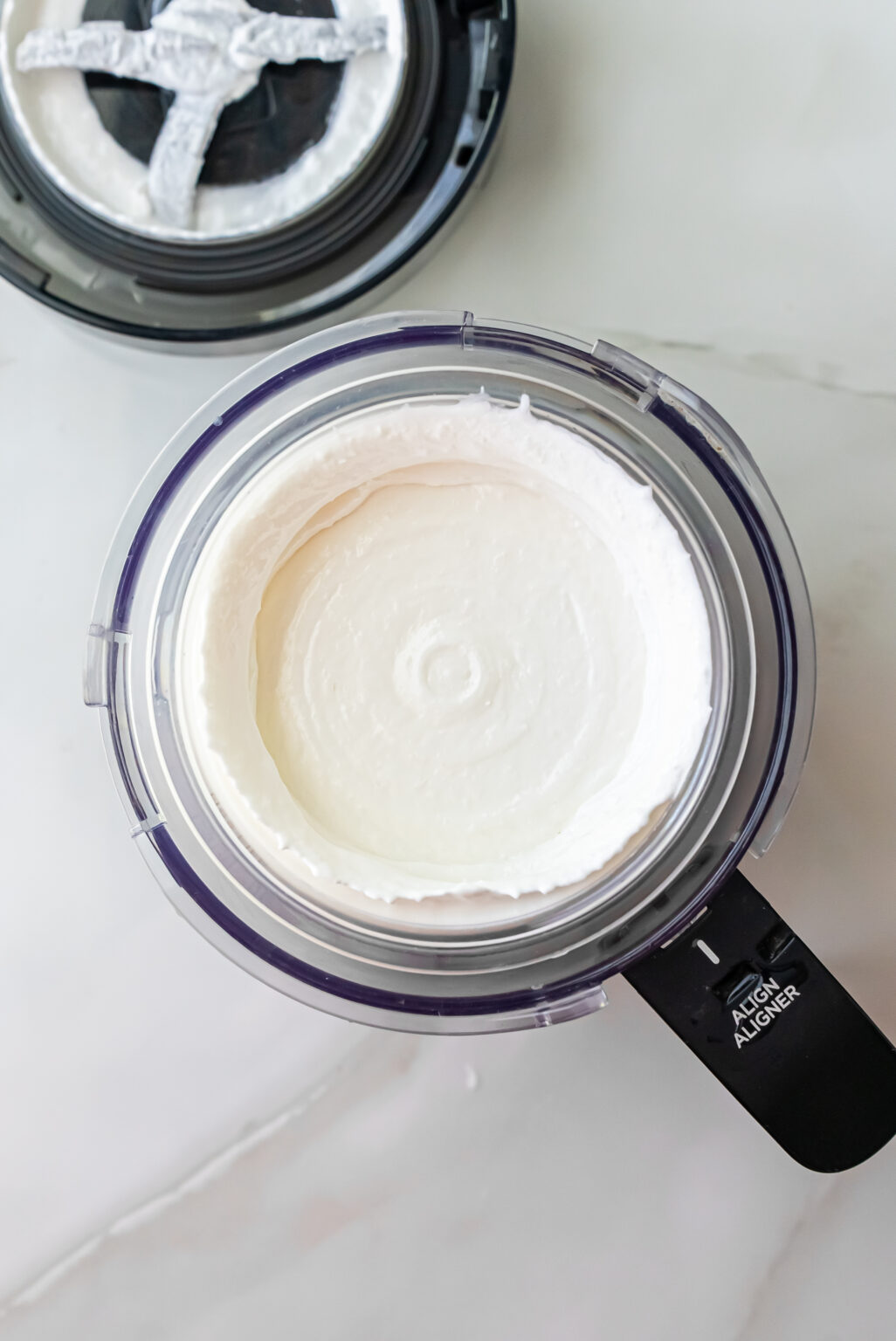 Top-down view of churned white ice cream in a Ninja Creami pint showing a smooth swirl pattern after the initial spin cycle for Lemonades Ninja Creami Ice Cream. The black lid sits beside it.