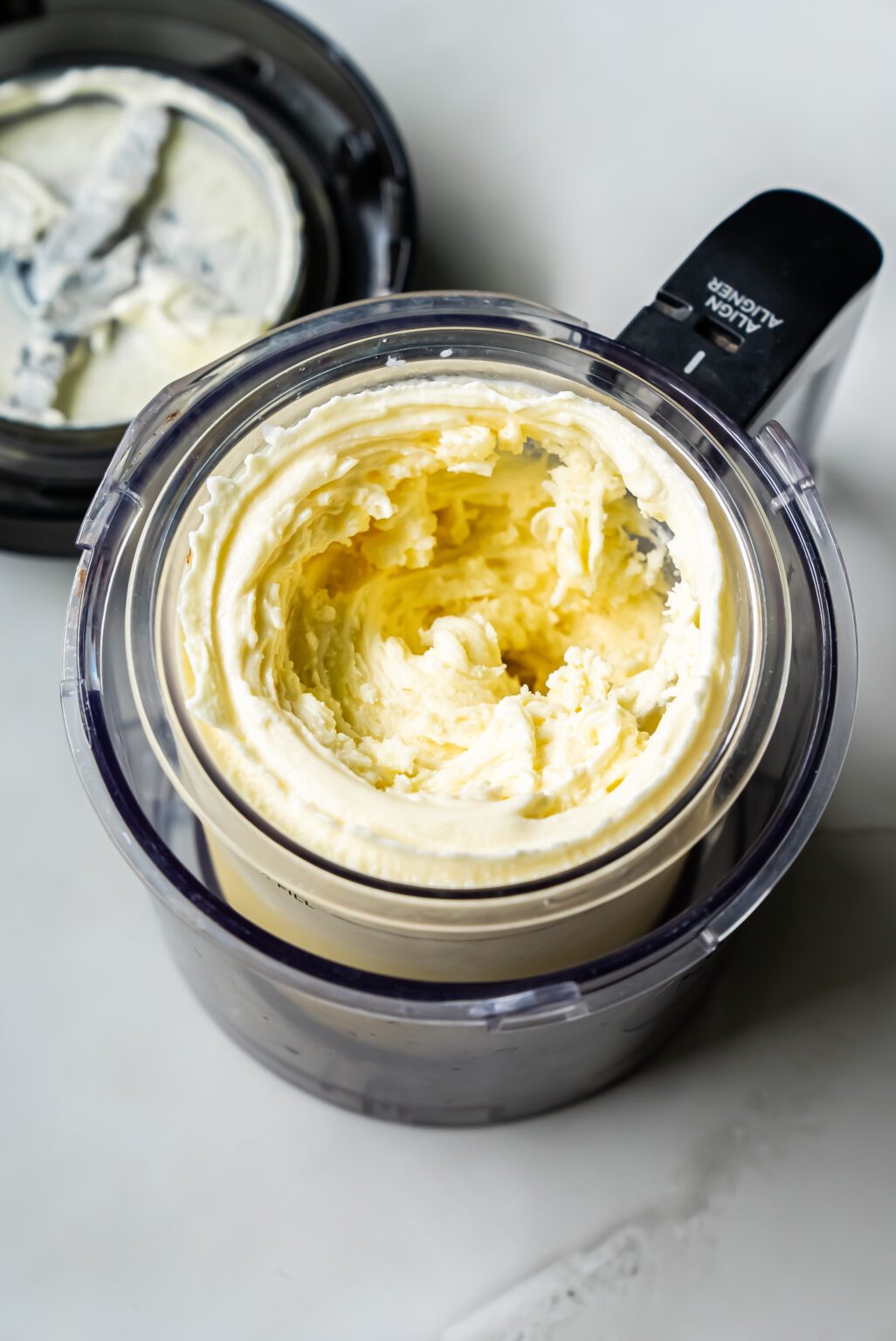 A top view of a Ninja Creami food processor containing freshly churned butter. The lid has been removed and is placed beside the processor on a white surface.