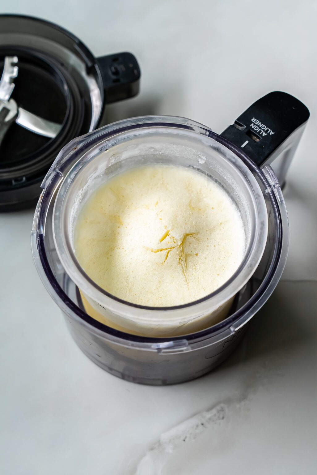 A food processor bowl is filled with partially blended ingredients for low carb delights. The lid of the processor, perhaps from a Ninja Creami, is set aside on a white surface.