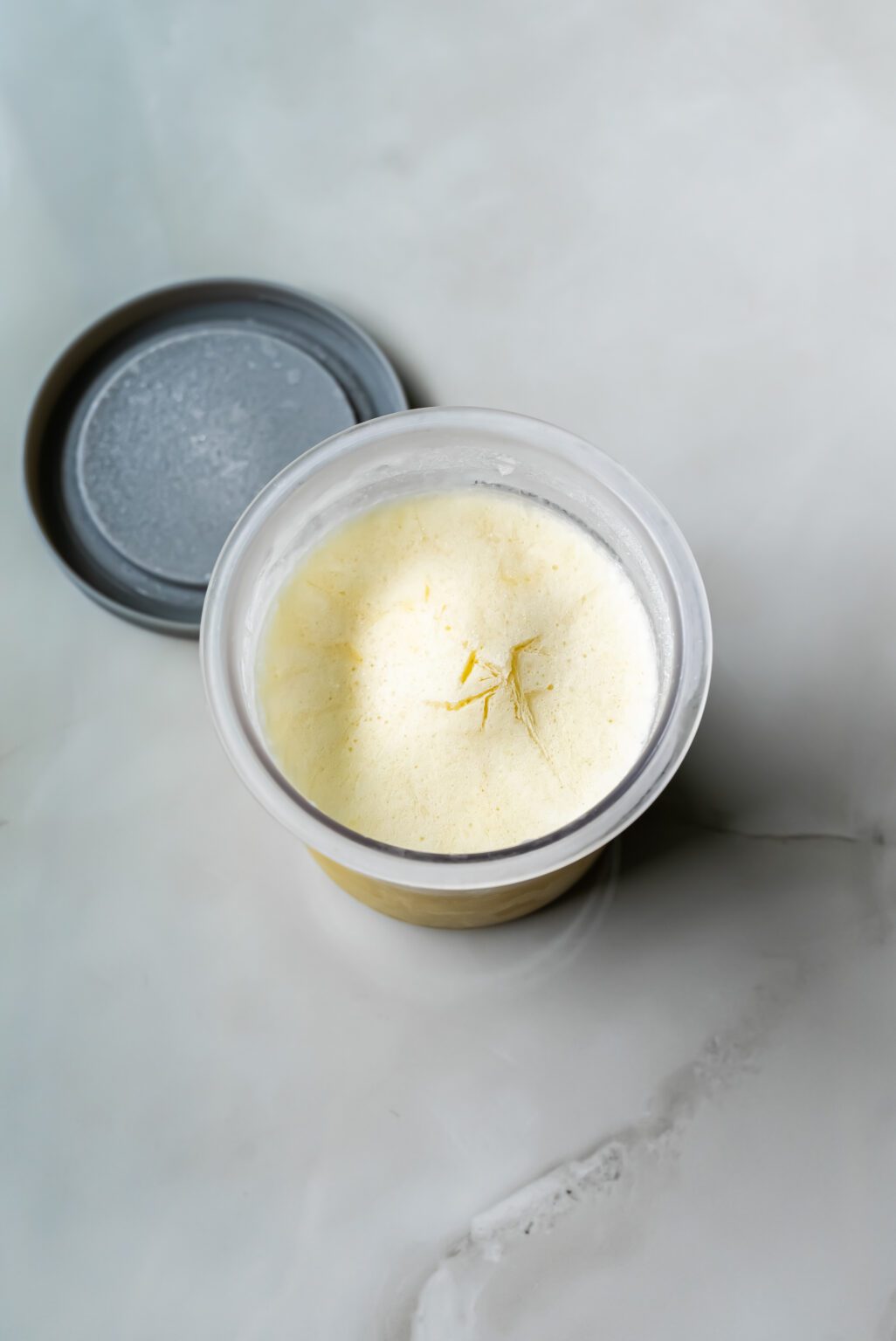 A clear container with a grey lid holds a pale, creamy low-carb substance with a few yellow strands on top, placed on a smooth, light-colored surface.
