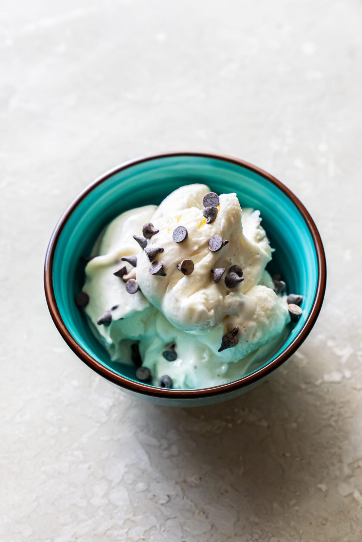 Top-down view of scoops of Mint Chocolate Protein Ice Cream served in a teal ceramic bowl with a brown rim. The white ice cream is generously topped with mini chocolate chips against a light marble background.