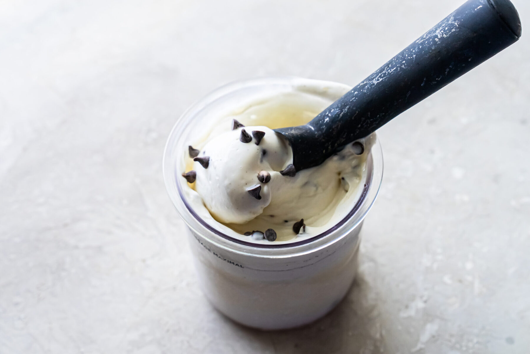 Angled overhead shot of a black scoop resting in a clear Ninja Creami pint filled with white Mint Chocolate Protein Ice Cream. Chocolate chips are visible on the surface and embedded within the frozen dessert.